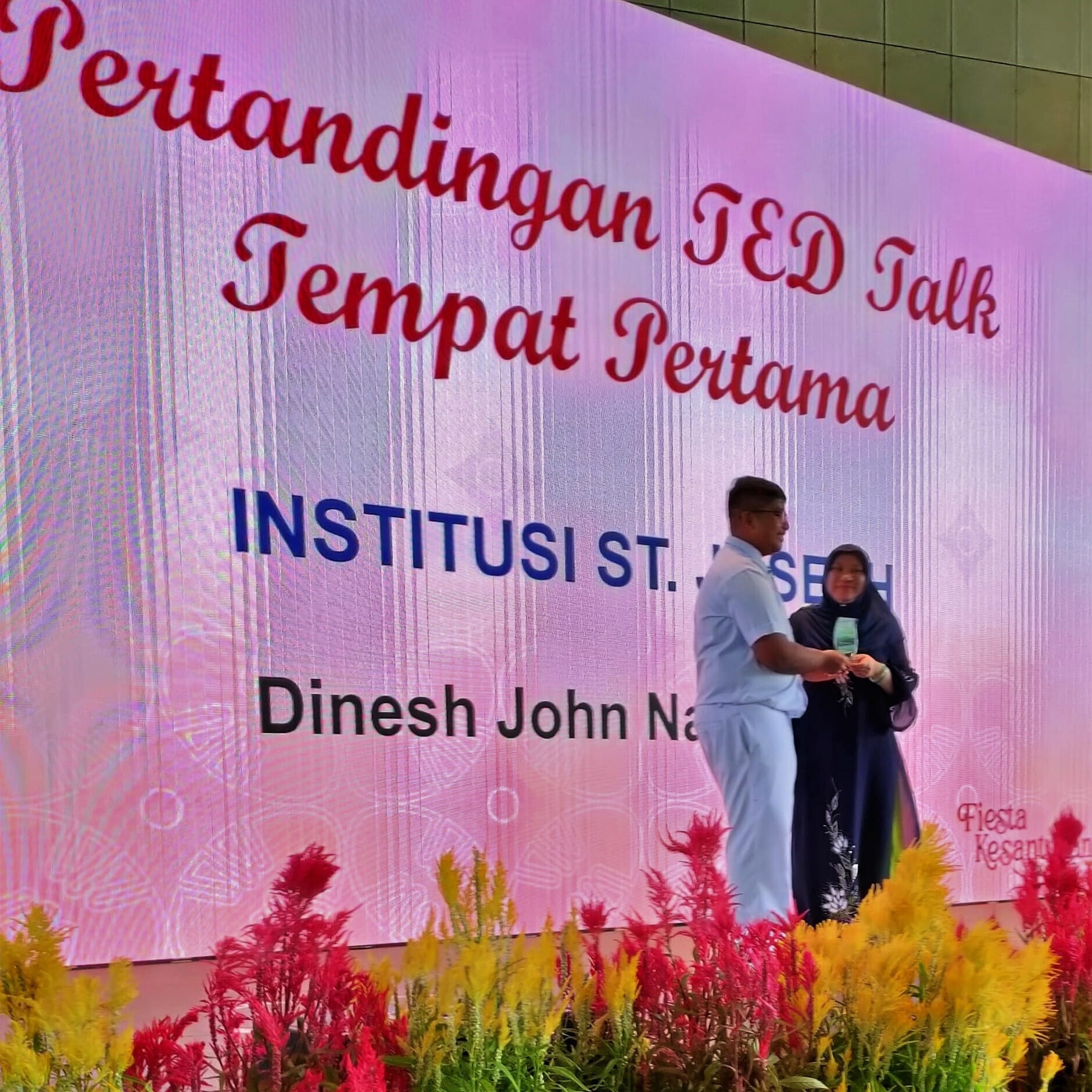 A man and woman exchange an award before a Malay-language screen reading 'TED Talk Competition First Place, Dinesh John'.